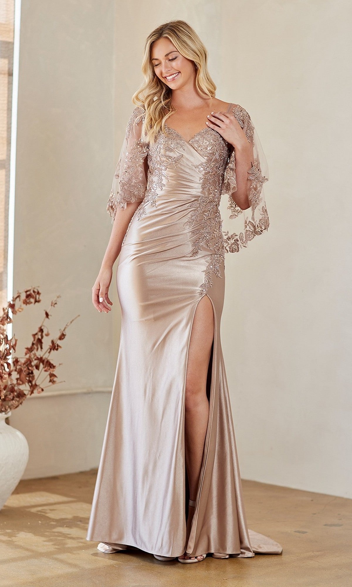 A woman in the Amelia Couture 8073 long formal dress with champagne lace embroidery, sheer sleeves, and a thigh-high slit stands indoors, smiling while looking down.