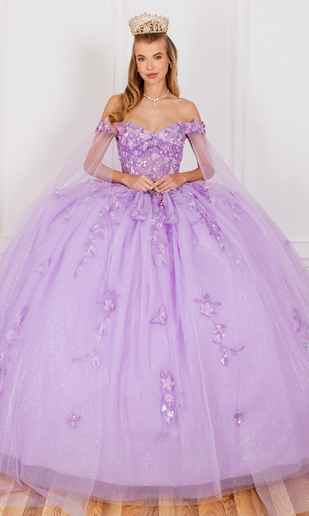 A woman in the Cinderella Couture 8075J Quinceanera Dress, featuring sparkly lavender fabric, floral details, and 3D appliques, accessorized with a gold crown and pearl necklace, stands against a white wall.