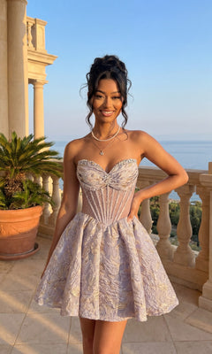 A woman in a lilac strapless Short Homecoming Dress (Amelia Couture 8075S) with a corset bodice stands on a stone balcony with plants and columns, gazing at the ocean.