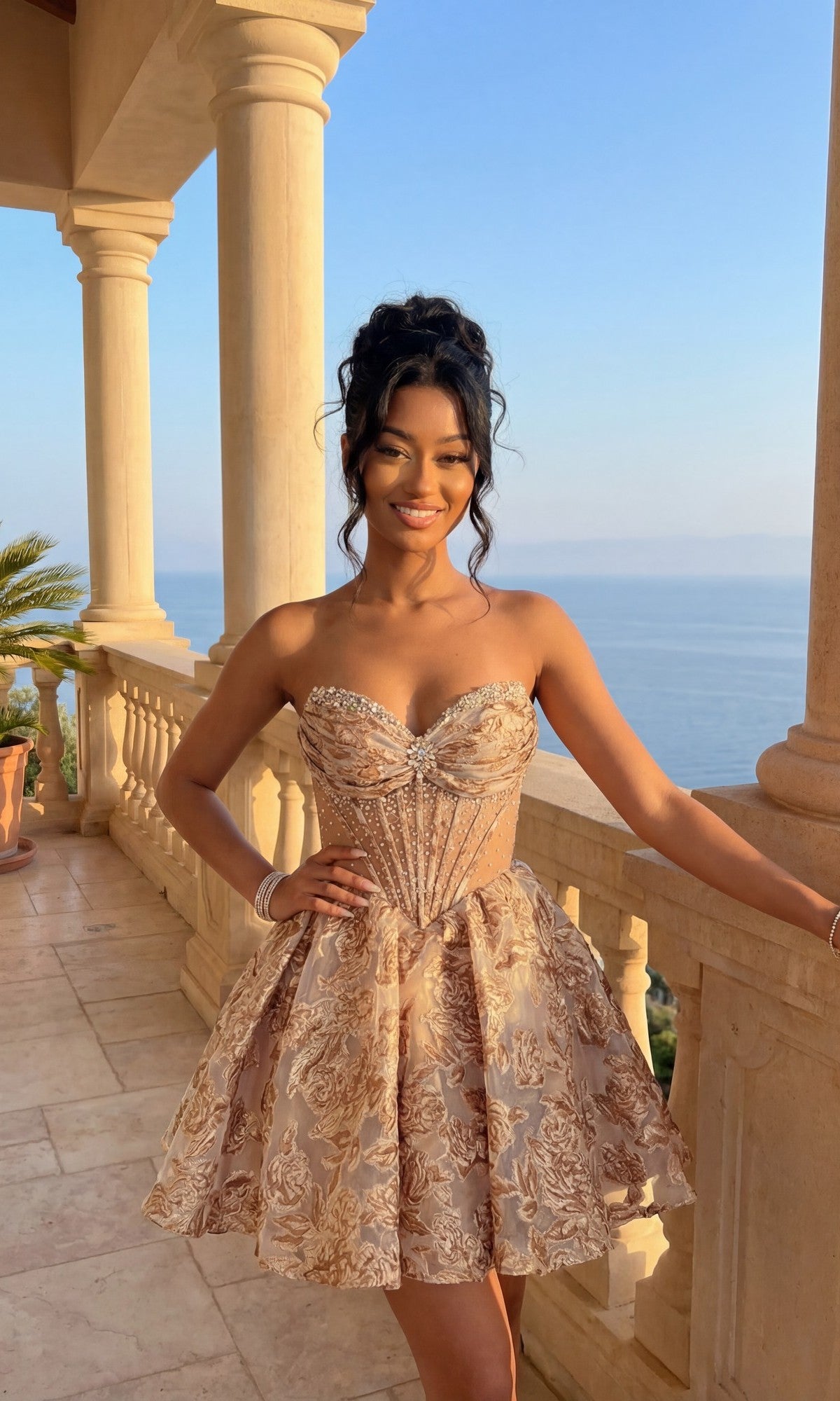 A woman in the mocha Short Homecoming Dress Amelia Couture 8075S, featuring a strapless corset bodice and floral brocade, stands smiling on a balcony overlooking the sea with one arm resting on the railing.