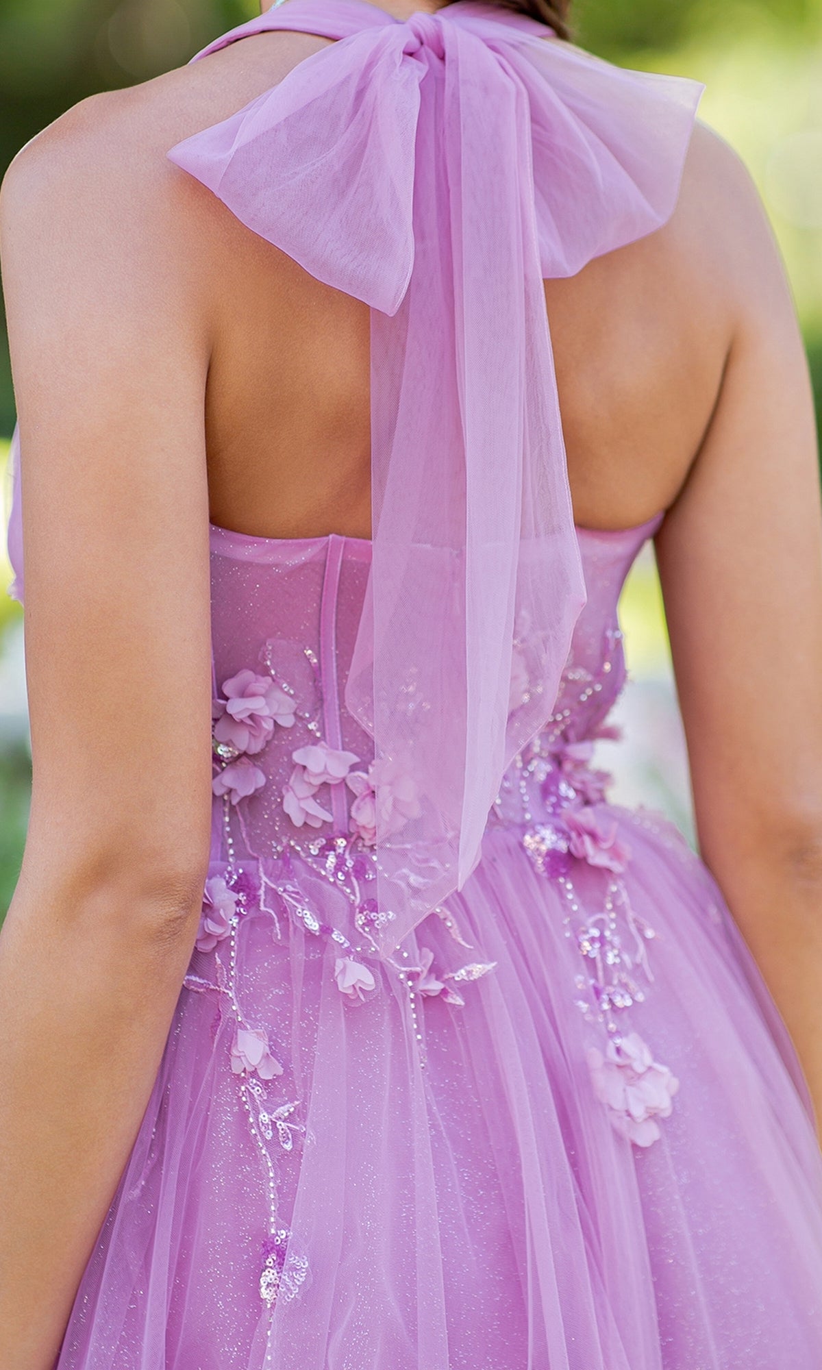 A person is seen from the back wearing the Cinderella Couture 8076J, a long halter prom dress in light purple with floral embellishments and a large neck bow.