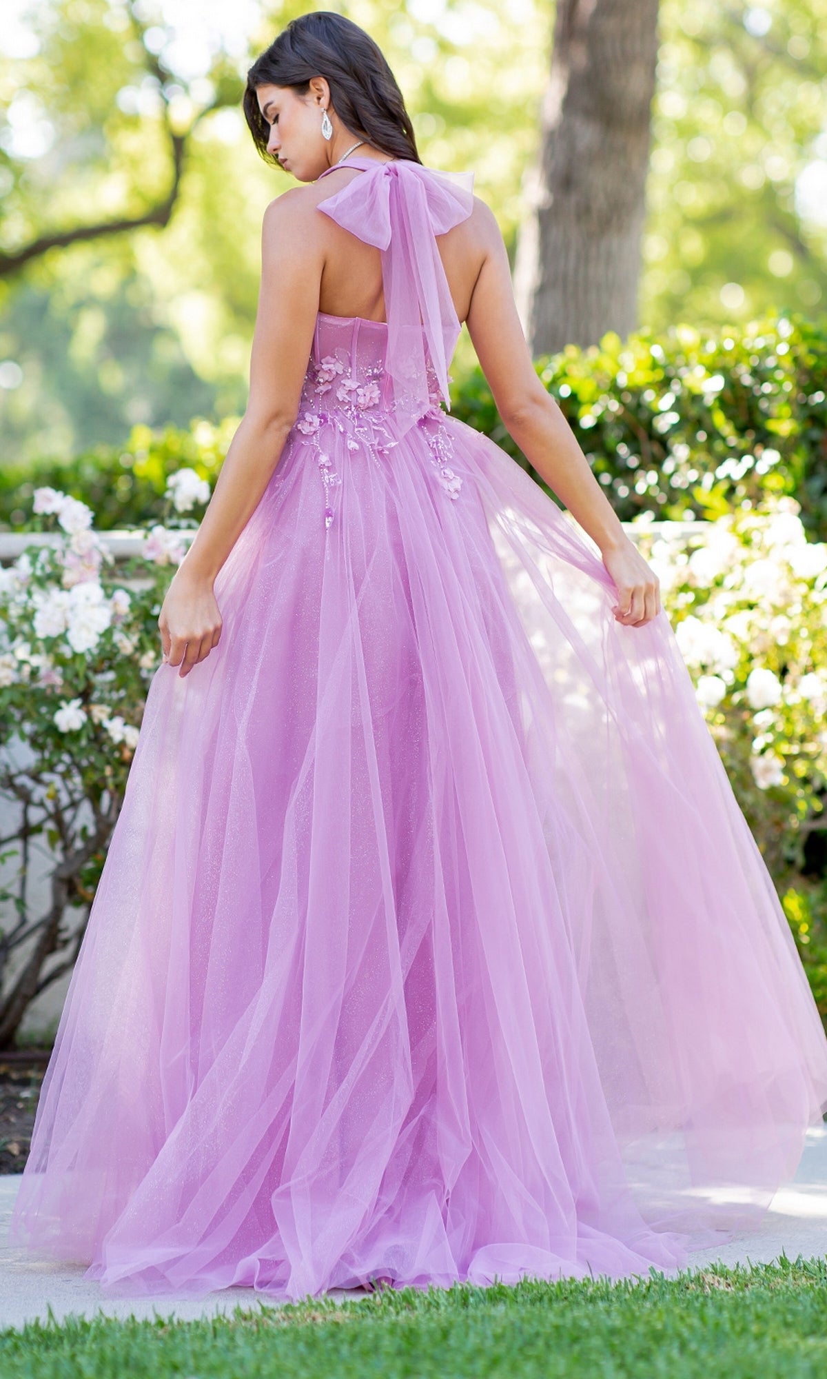 A woman stands outdoors, facing away, wearing the Cinderella Couture 8076J Long Halter Prom Dress with floral details and flowing pink tulle, holding the sides of her gown.
