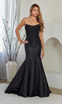 A woman poses indoors with her hand on her hip wearing the black Amelia Couture 8080 long prom dress, featuring a strapless embellished corset bodice and fit-and-flare skirt, against a neutral background with bare branches.