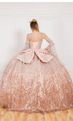 A woman in the Quinceanera Ball Gown 8080J—sparkling pink ombre with silver details and a large bow—faces a white ornate wall, crowned in gold.