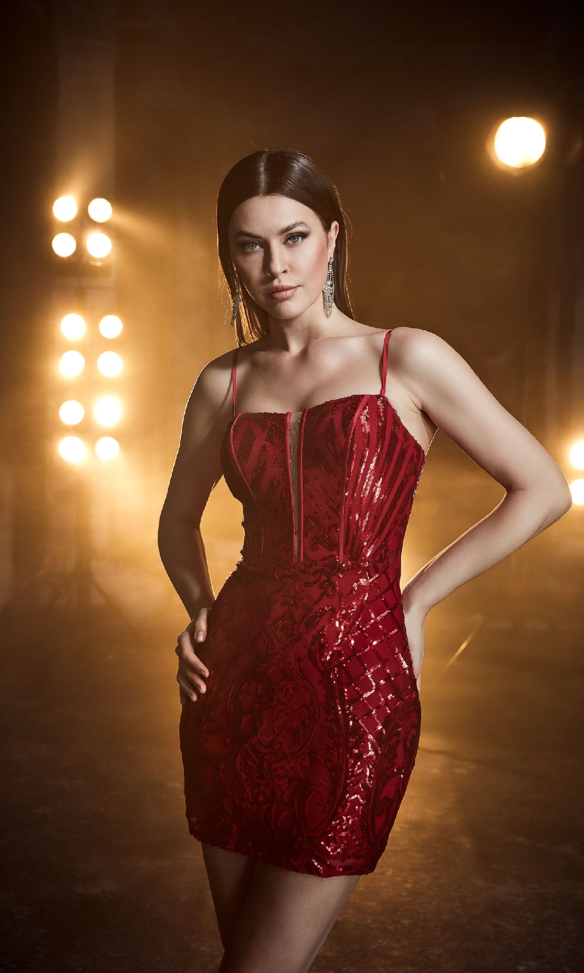 A woman stands confidently under bright stage lights in a dim room, wearing the Red Sequin Homecoming Dress: Aerie Couture 808.