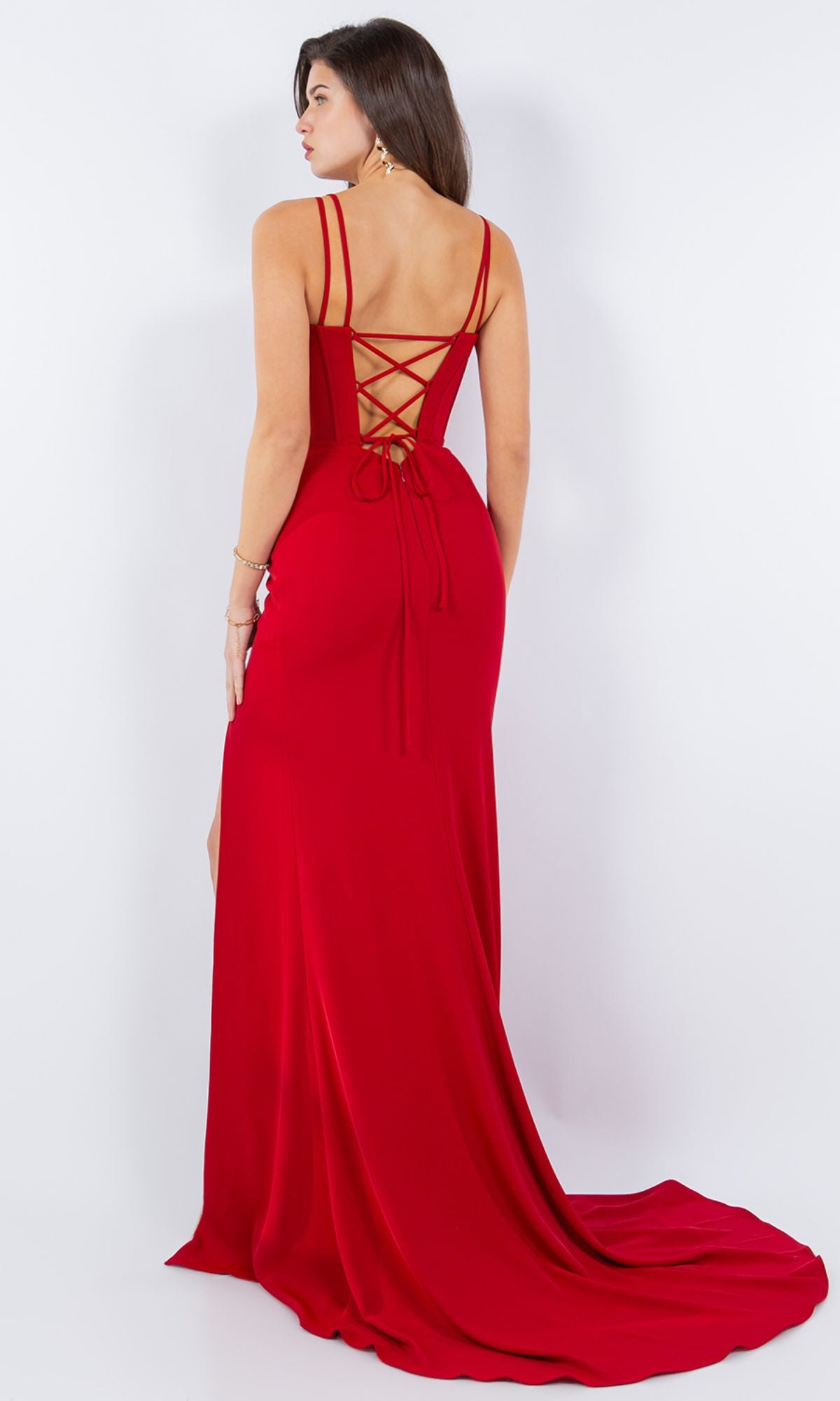 A woman in the Cinderella Couture 8091J Strappy Corset Prom Dress, floor-length and red with pleats and a lace-up back, stands facing away, glancing over her shoulder.
