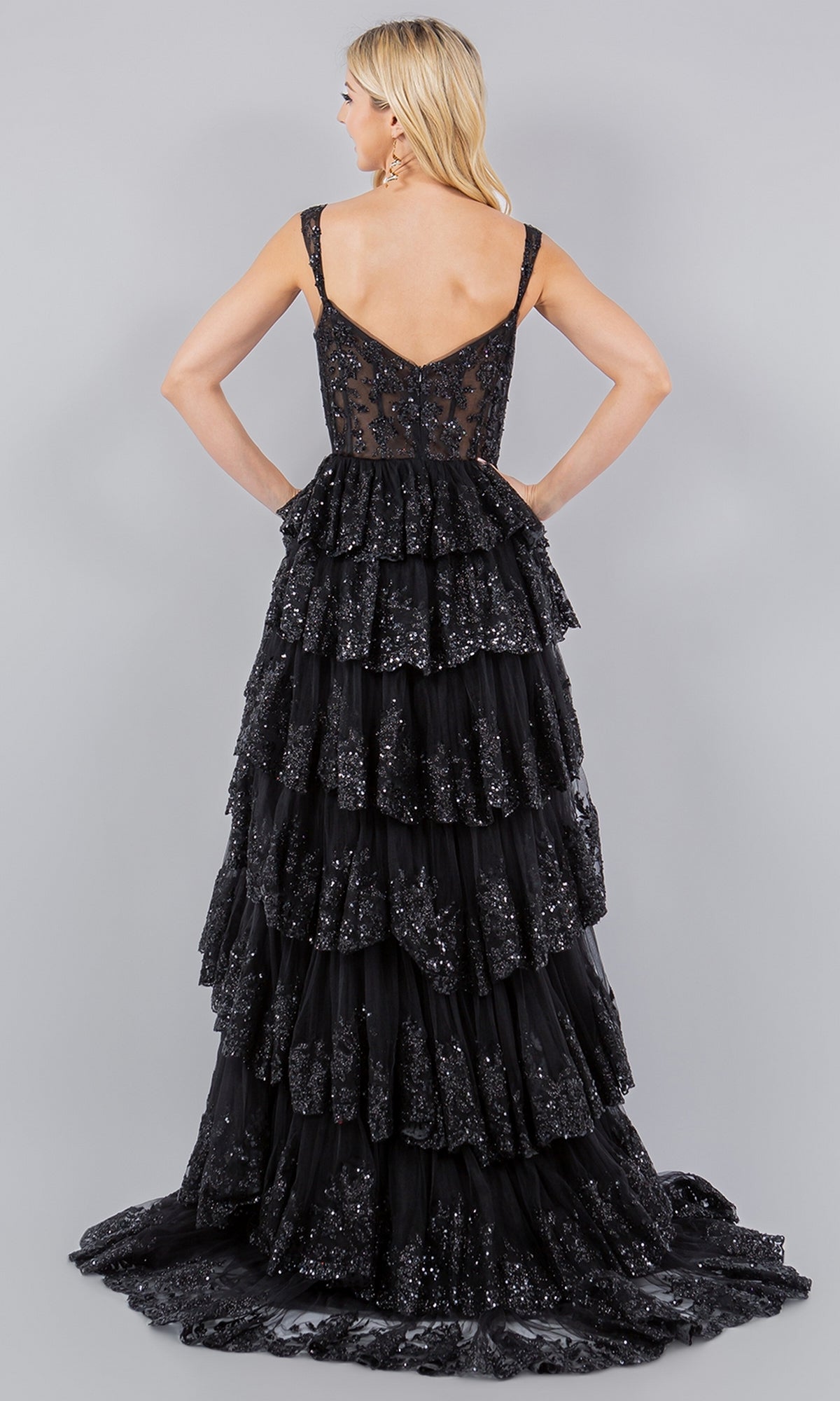 A woman stands facing away in the Cinderella Couture 8093J—a black, sleeveless, floor-length prom dress with layered ruffles and shimmering sequins—set against a plain gray background.
