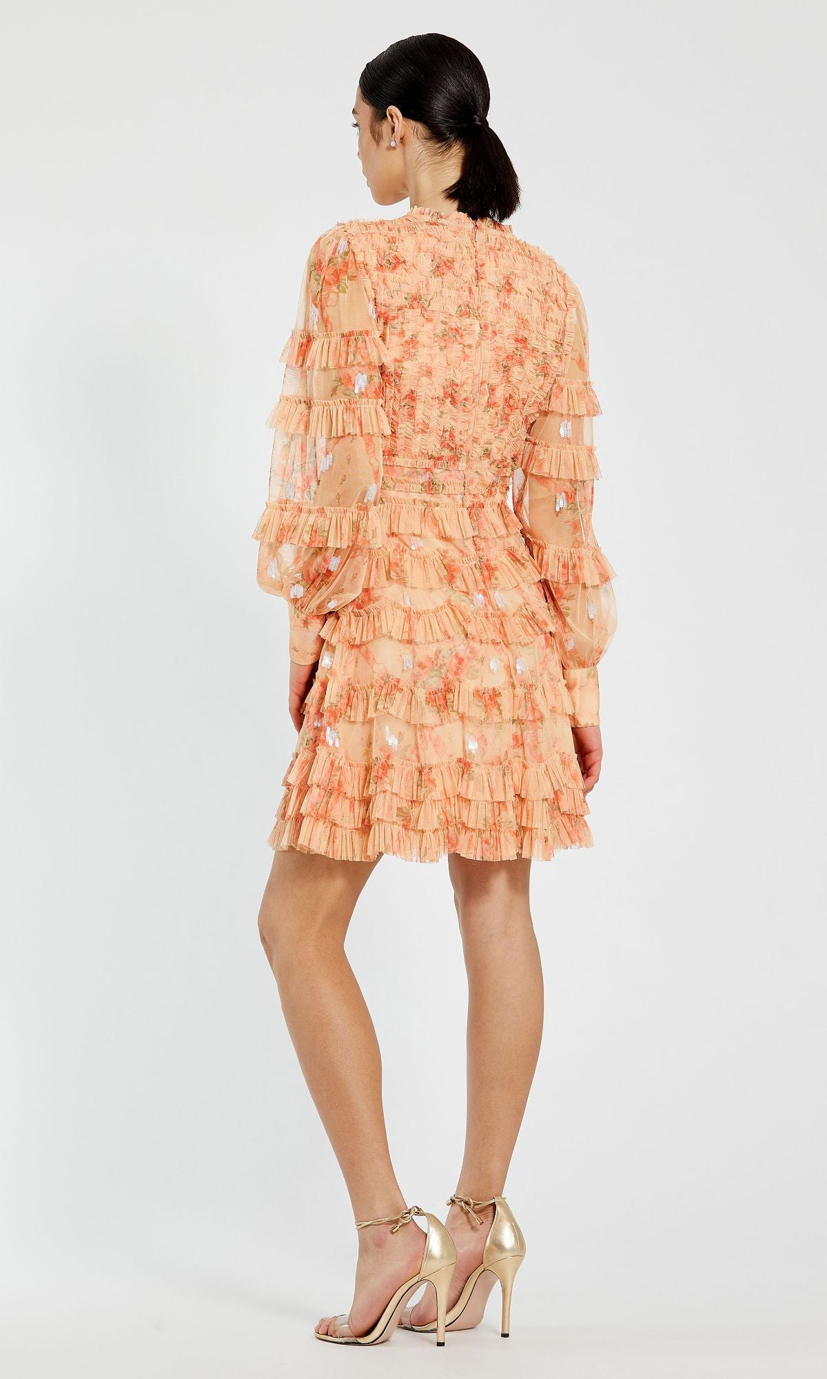 A woman stands facing away in the Mac Duggal 8094, a peach floral wedding guest dress featuring long sleeves, delicate ruffles, and gold high-heeled sandals—a perfect choice for special occasions.