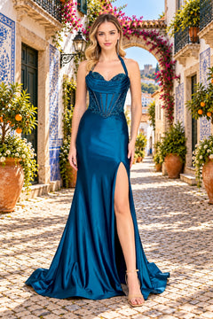 A woman stands on a cobblestone street wearing an Amelia Couture 8096 long prom dress, a floor-length, deep blue satin corset gown with a beaded bodice and a high side slit.