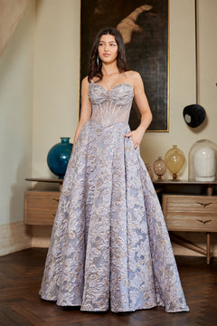 A woman wearing the Long Prom Dress: Amelia Couture 8098, a strapless floral gown with a sweetheart neckline, a sheer corset-style bodice, and a voluminous skirt featuring a metallic jacquard pattern.