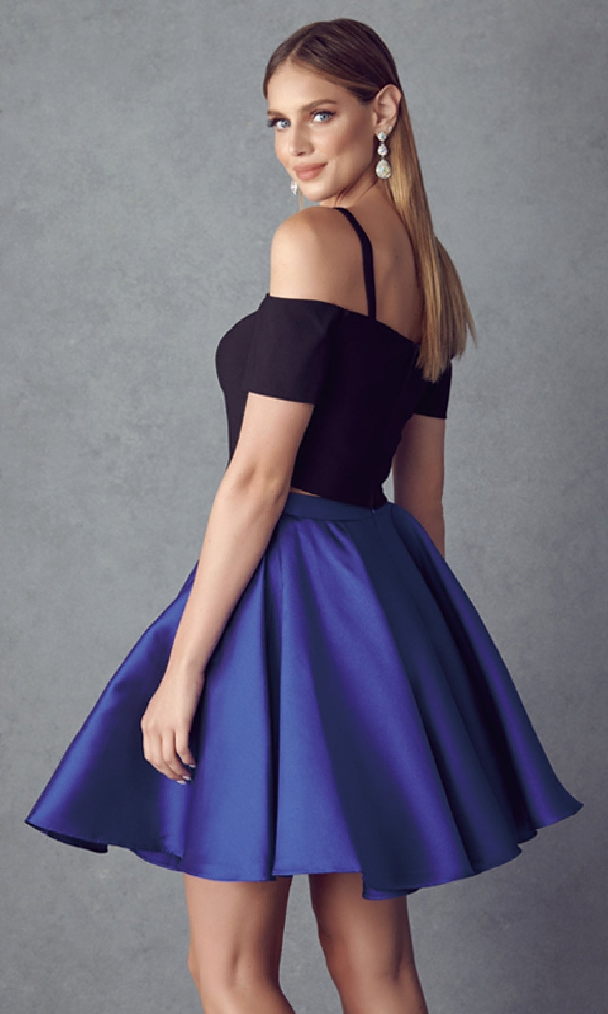 A woman models the Juliet 809 Short Party Dress, featuring a black off-the-shoulder top and a flared royal blue A-line skirt, standing against a plain gray background.