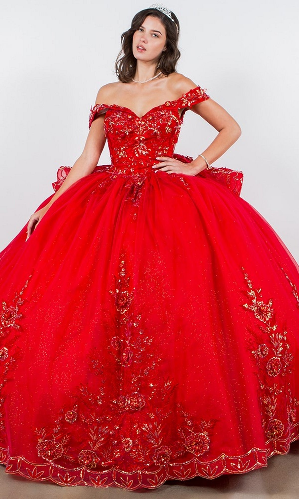 A woman in the Quinceanera Ball Gown 8100J—bright red, off the shoulder, with gold floral embroidery, beaded details, a full skirt, lace up back—stands against a plain background wearing a sparkling tiara.