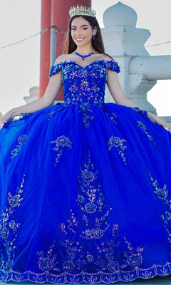 Young woman outdoors in the Quinceanera Ball Gown 8100J with off-the-shoulder design, iridescent beaded details, floral embroidery, lace-up back, and royal blue skirt, smiling as she holds out her dress.