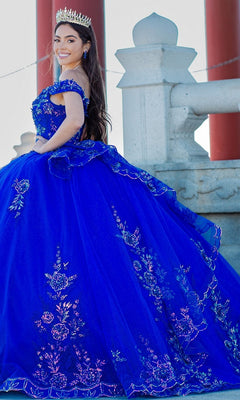 A woman in the Quinceanera Ball Gown 8100J, featuring an off-the-shoulder design and iridescent beaded details, smiles and poses outdoors beside a stone railing while wearing a crown.