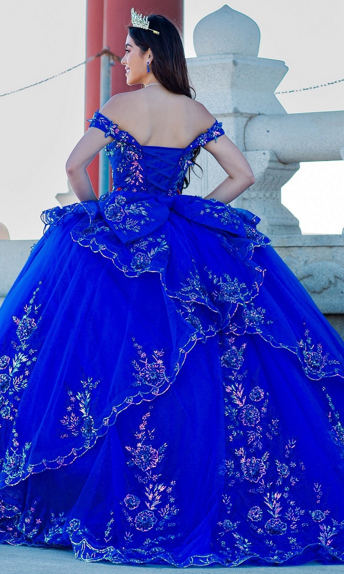 Wearing the Quinceanera Ball Gown 8100J in bright blue with iridescent beads and a tiara, a person stands outdoors with their back to the camera.