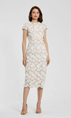 A woman stands against a plain background wearing the Mac Duggal 81022 Midi Lace Cocktail Dress in white with short sleeves, floral lace pattern, and nude heels.