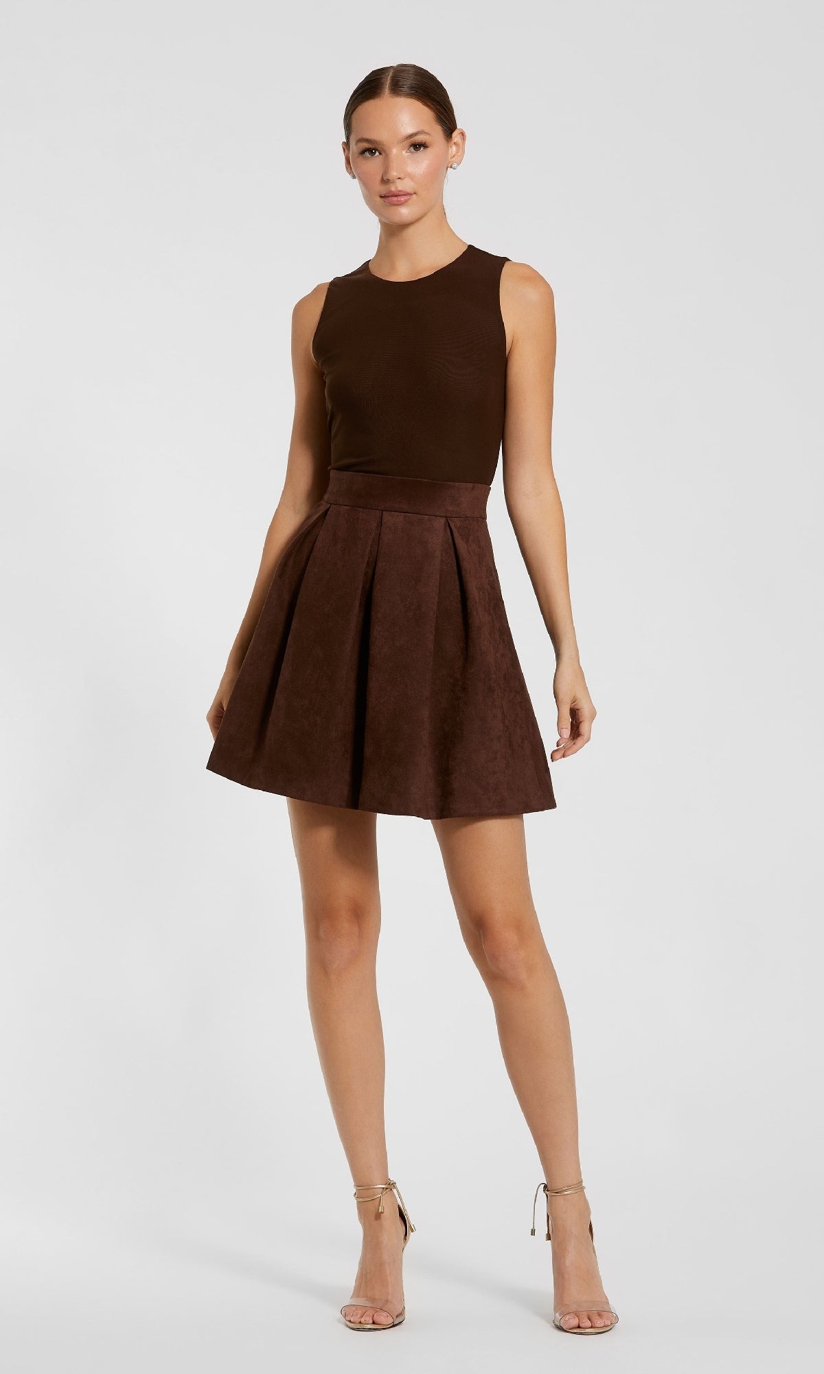 A woman poses against a plain background in the Mac Duggal 81071 short brown wedding-guest dress, featuring a sleeveless top and high-waisted pleated skirt, paired with beige high-heeled sandals.