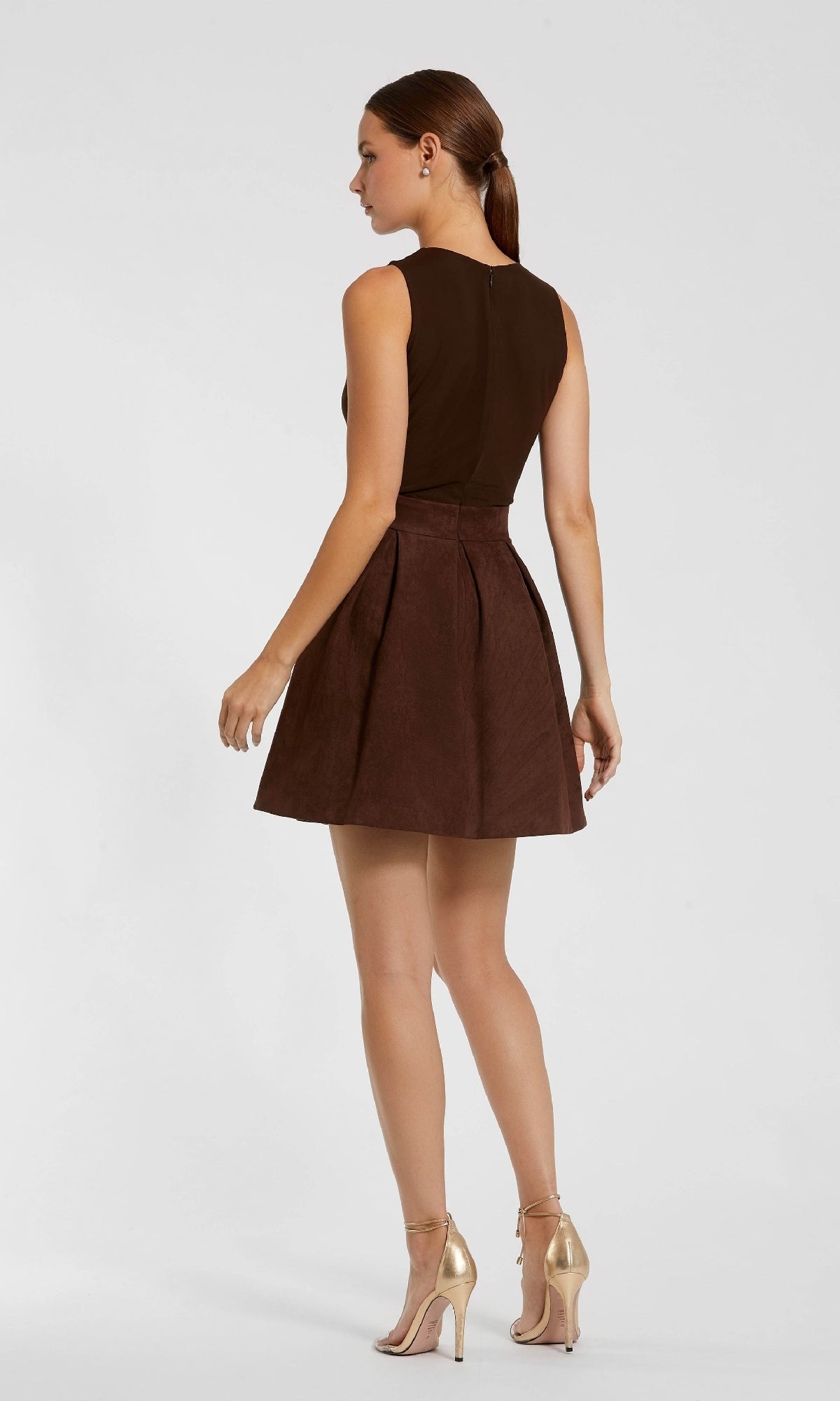 A woman faces away, wearing the Mac Duggal 81071 short brown wedding-guest dress with pleats, paired with gold high-heeled shoes.