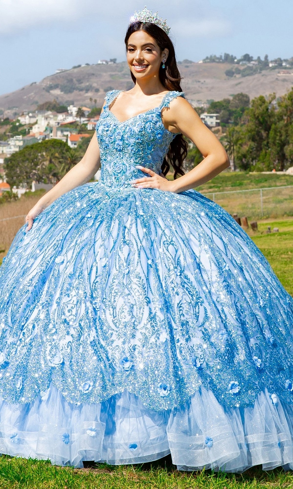 A woman in the Quinceanera Ball Gown 8110J, featuring a sparkling blue skirt and embroidered bodice with a tiara, stands on grass with hills and houses in the background.