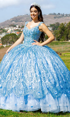 A woman in the Quinceanera Ball Gown 8110J, featuring a sparkling blue skirt and embroidered bodice with a tiara, stands on grass with hills and houses in the background.