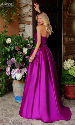 A woman wearing the Clarisse Designs 811167 Long Prom Dress, featuring a strapless bodice and detachable overskirt, stands with her back to the camera near a dark doorway, surrounded by potted flowers and stone walls.