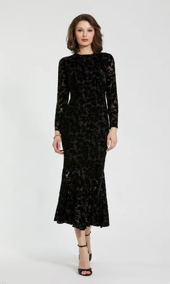 A woman stands against a plain background, wearing the Mac Duggal 81129 midi party dress in black, featuring burnout velvet fabric with long sleeves and floral patterns, styled chicly with black heeled sandals.