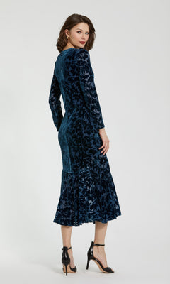 A woman stands against a plain light background, angled slightly away, wearing the Mac Duggal 81129—a fitted, long-sleeve blue burnout velvet midi party dress with a dark floral pattern and flared hem—styled with black ankle-strap heels for an elegant look.