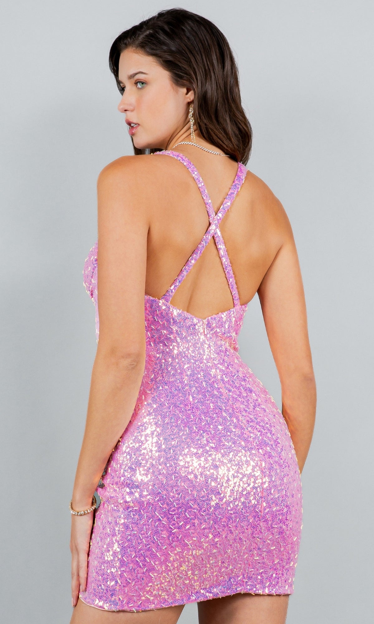 A woman poses with her back to the camera, highlighting the Short Sweetheart Sequin Homecoming Dress 8113J in sparkly pink with crisscross straps, set against a plain gray background.