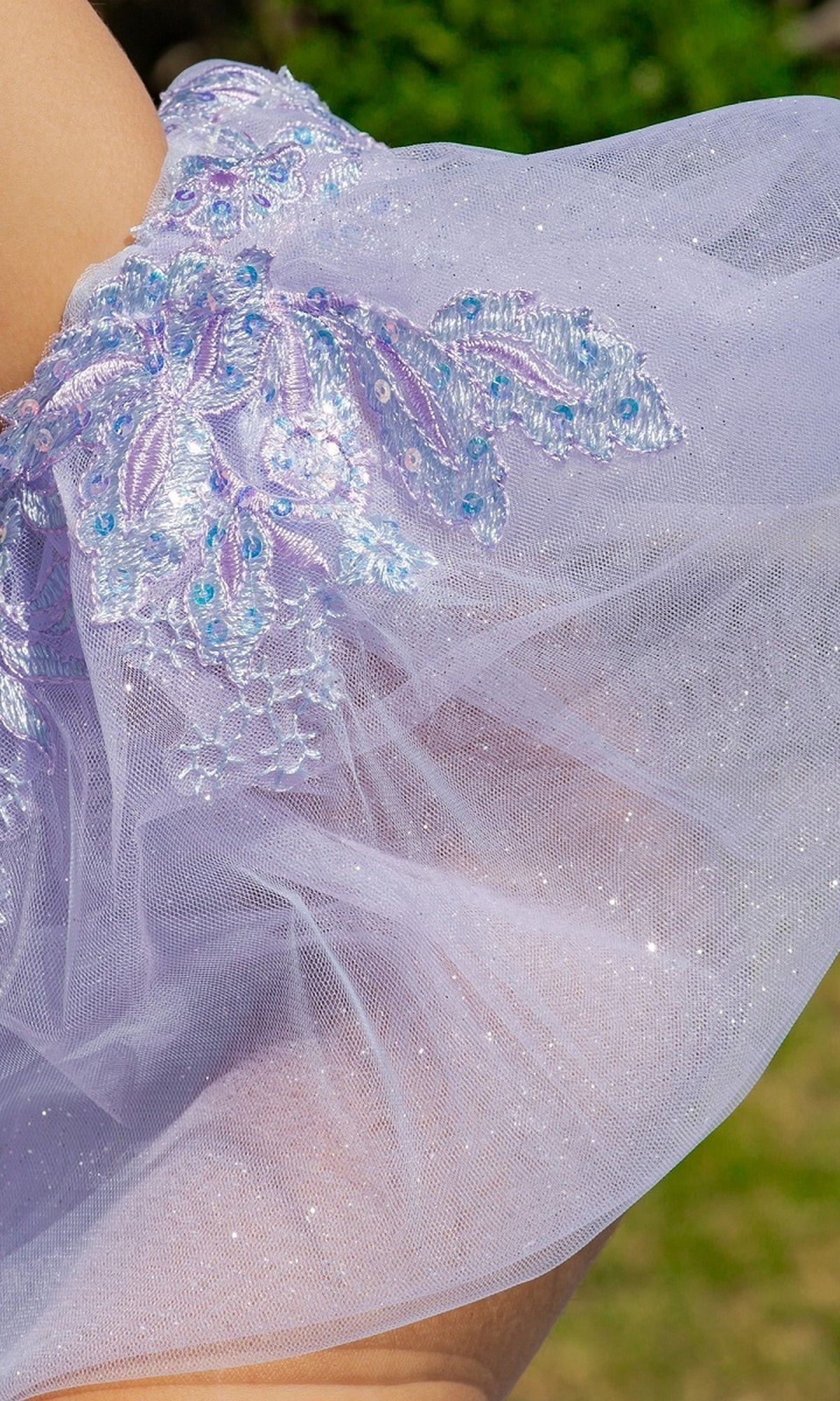 Close-up of Quinceanera Ball Gown 8115J showing sheer light purple fabric with embroidered floral patterns and sequins against a blurred green background.