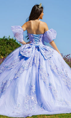 A woman stands outdoors facing away, wearing the Quinceanera Ball Gown 8115J in light purple with floral embellishments, removable puff sleeves, and a lace-up back.