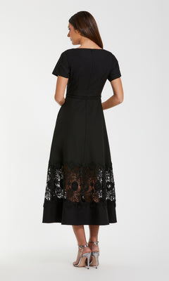 A woman, seen from behind, wears the Mac Duggal 81206 Short Sleeve Midi Cocktail Dress in black with a sheer lace hem panel and silver heels against a plain white background.