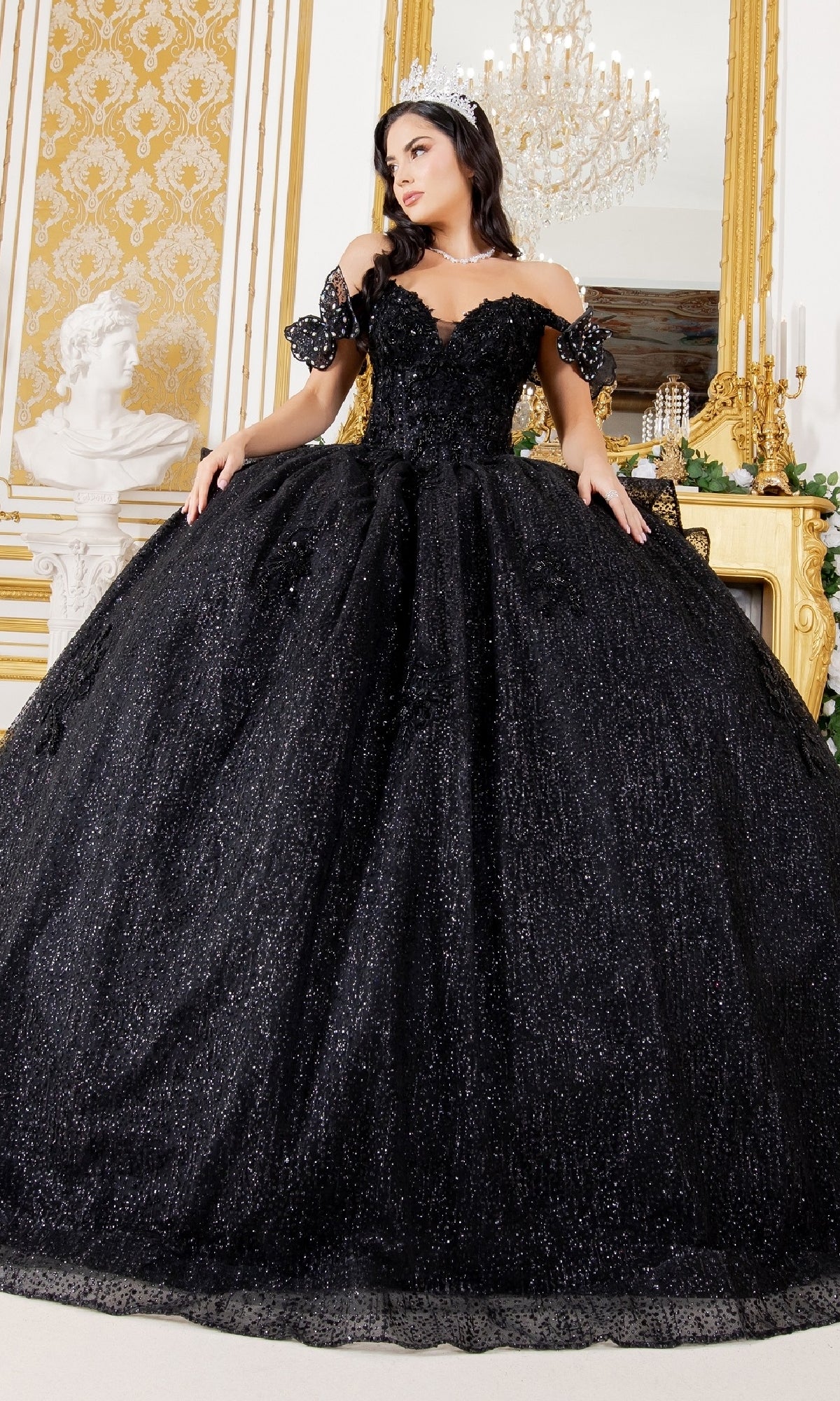 A woman in the Quinceanera Ball Gown 8120J, featuring a black off-the-shoulder design with butterfly back detail and a tiara, stands in an ornate room adorned with gold decor and a chandelier.