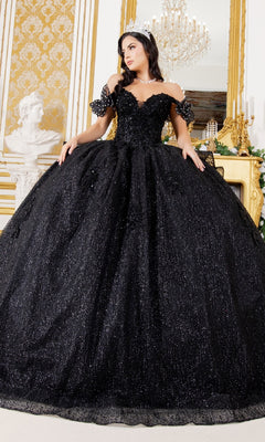 A woman in the Quinceanera Ball Gown 8120J, featuring a black off-the-shoulder design with butterfly back detail and a tiara, stands in an ornate room adorned with gold decor and a chandelier.