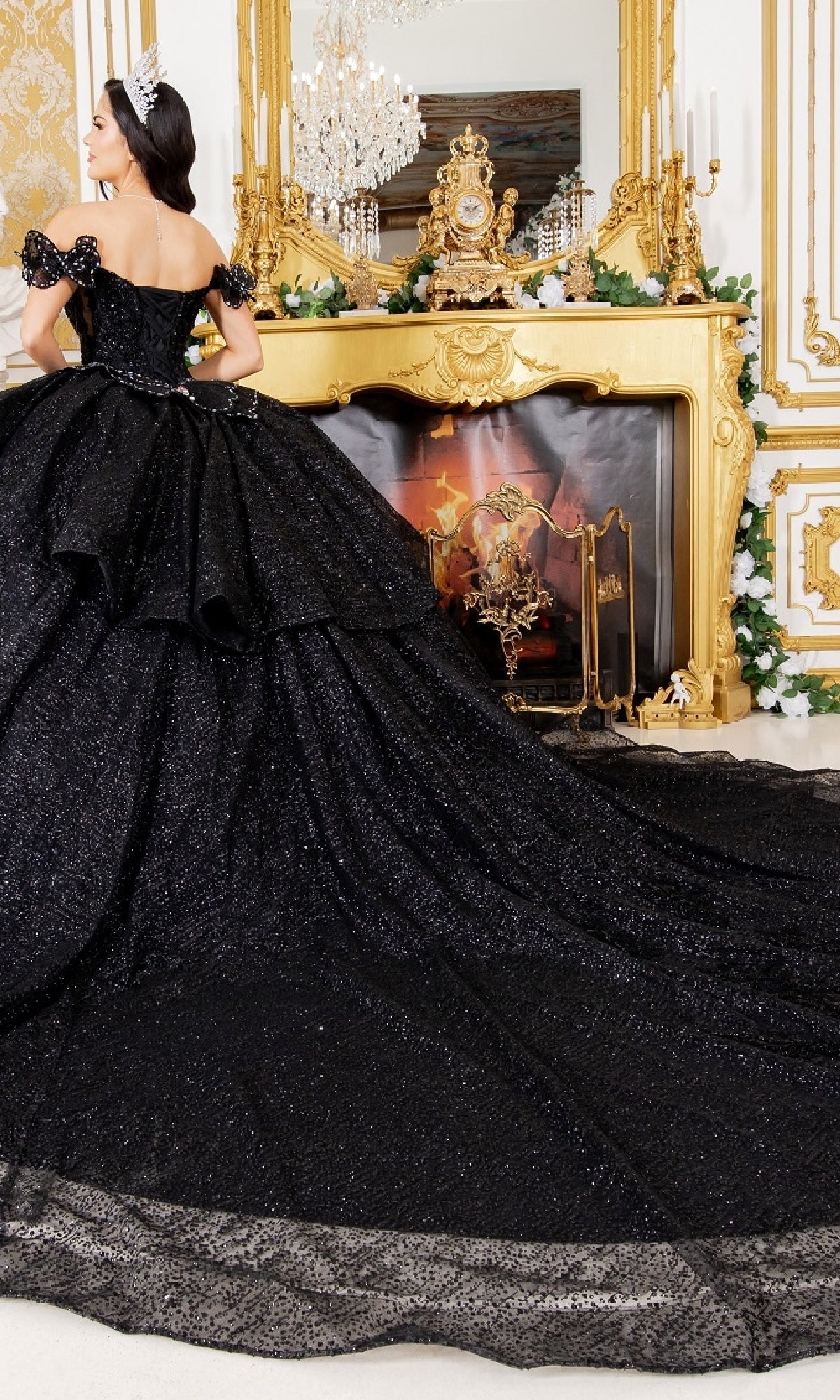 A woman wearing the Quinceanera Ball Gown 8120J, an off-the-shoulder black glitter ball gown with a long train, stands before an ornate gold fireplace adorned with greenery, flowers, and a large mirror.