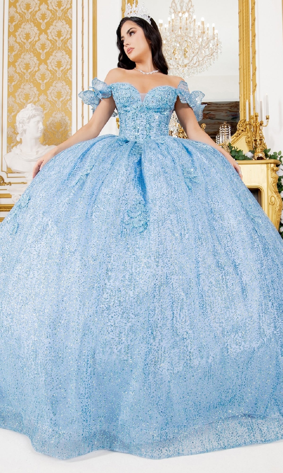 A woman poses in an ornate room wearing the Quinceanera Ball Gown 8120J, an off-the-shoulder sparkling light blue dress with delicate sleeves and a tiara.
