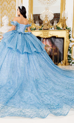 A woman in the Quinceanera Ball Gown 8120J, a sparkling blue dress with a long train and butterfly back detail, stands facing away from the camera in front of a gold mantelpiece.