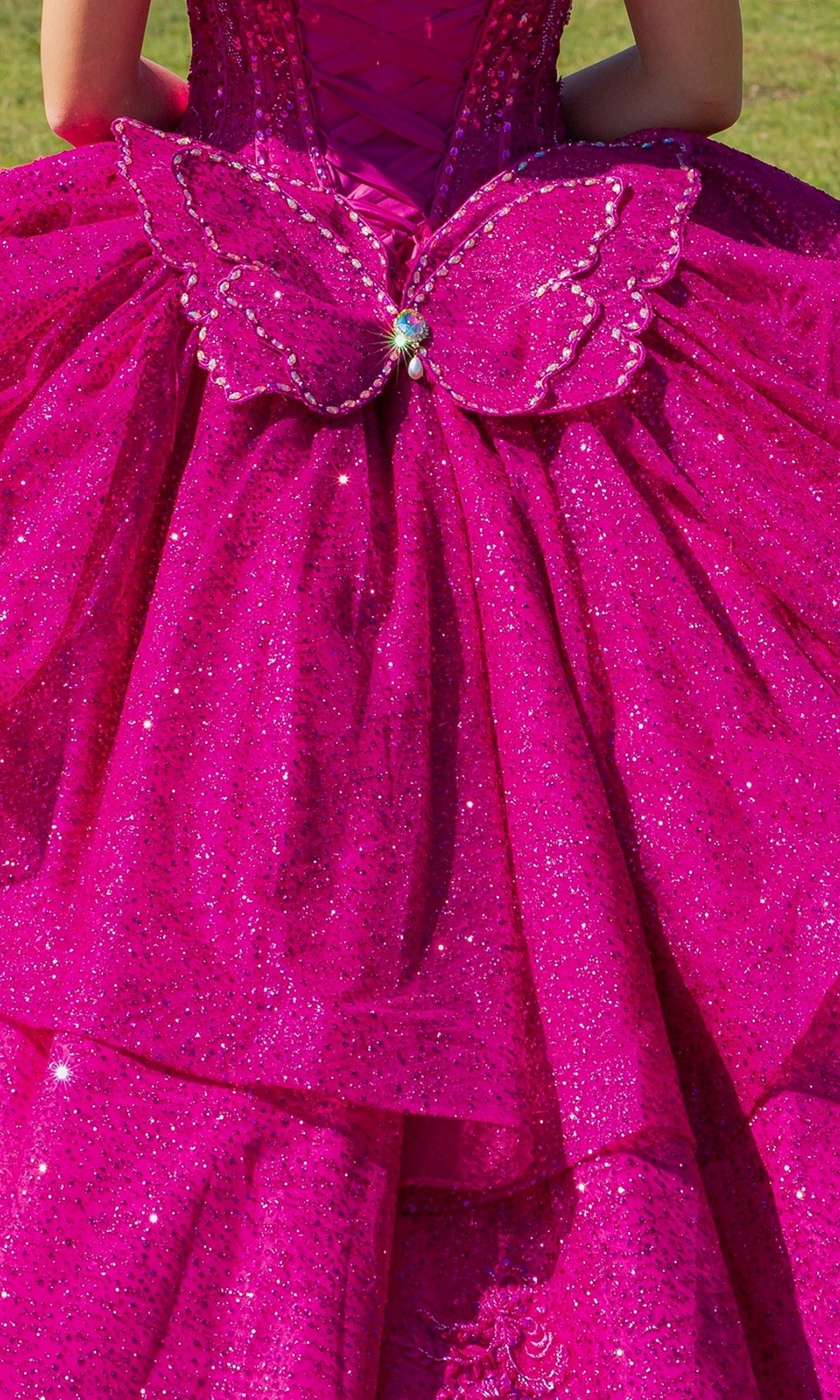 Wearing the Quinceanera Ball Gown 8120J in bright pink with glitter, an off-the-shoulder design, and butterfly back detail, a person stands gracefully on the grass.