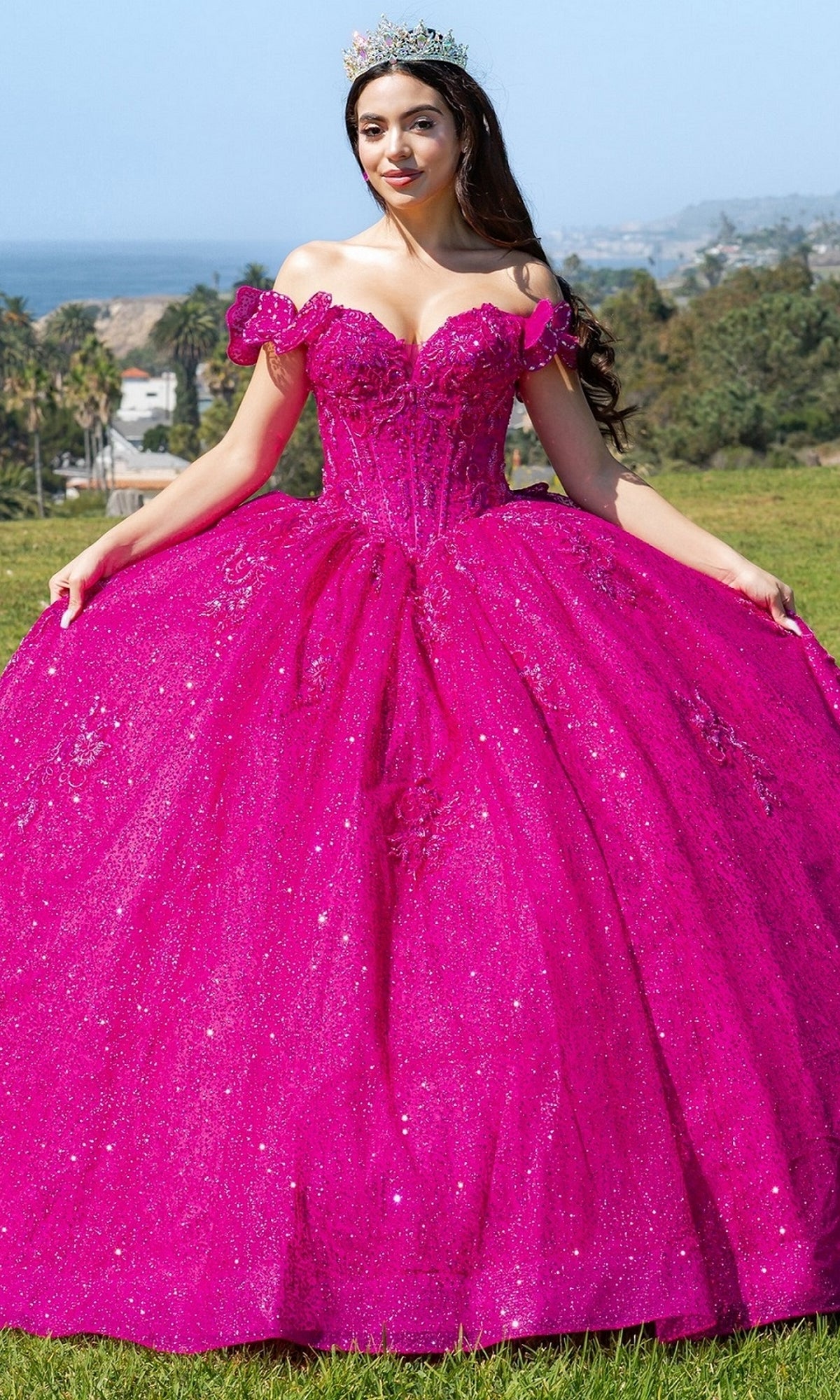 A woman in the sparkling bright pink Quinceanera Ball Gown 8120J with an off-the-shoulder design and a tiara stands outdoors on grass, framed by trees and hills.