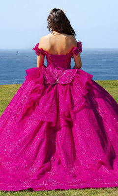 A woman in the Quinceanera Ball Gown 8120J, featuring a bright pink sparkle, off-the-shoulder sleeves, and butterfly back detail, stands on grass facing the ocean.