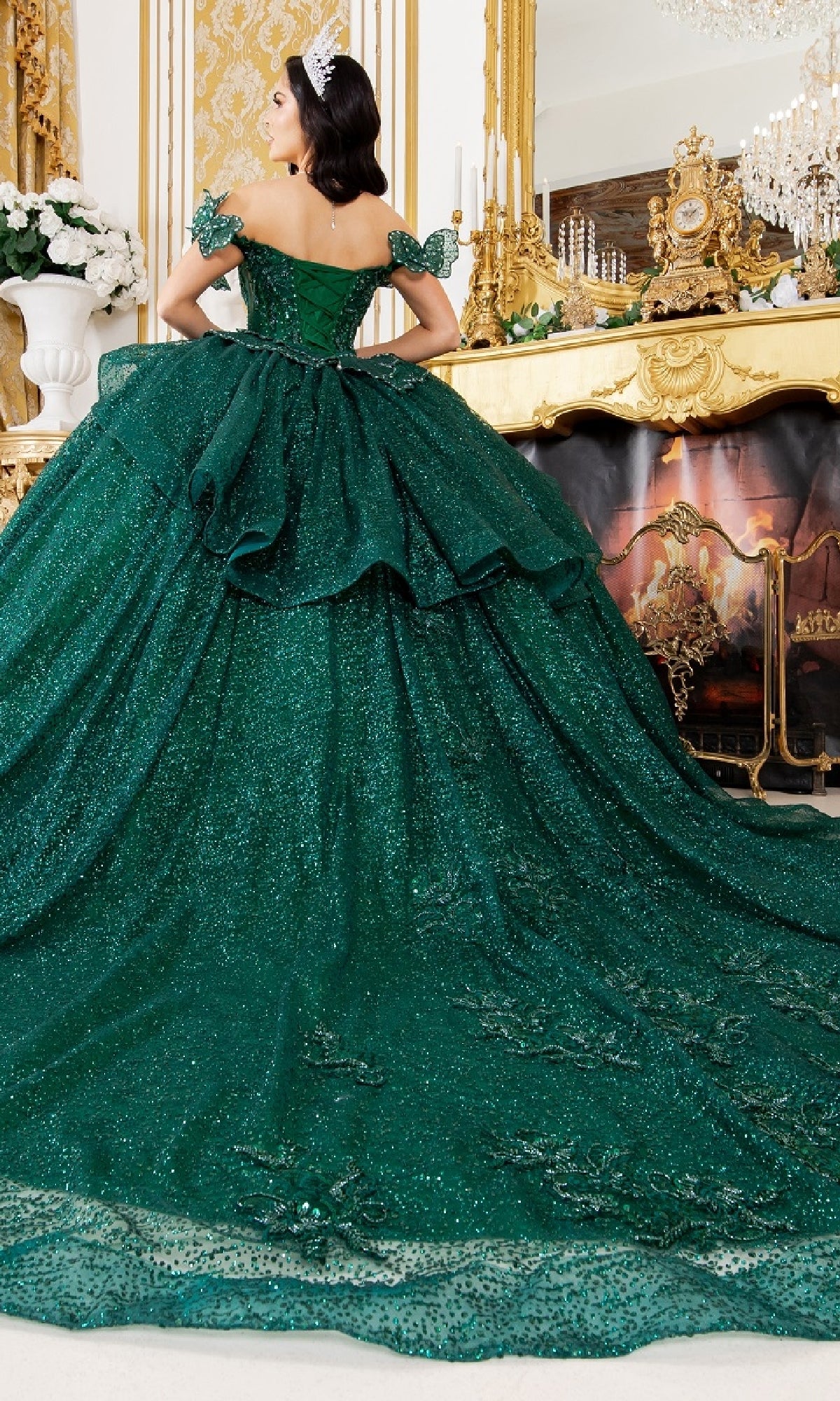 A woman wearing the Quinceanera Ball Gown 8120J, a sparkling dark green off-the-shoulder dress, stands in an ornate room with gold decor and a large fireplace, crowned with a tiara.
