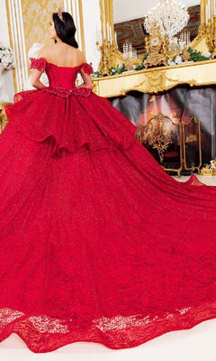 A woman in the Quinceanera Ball Gown 8120J—an off-the-shoulder, sparkling red gown with a long train and butterfly back—stands facing a gold ornate fireplace and chandelier, her back to the camera.