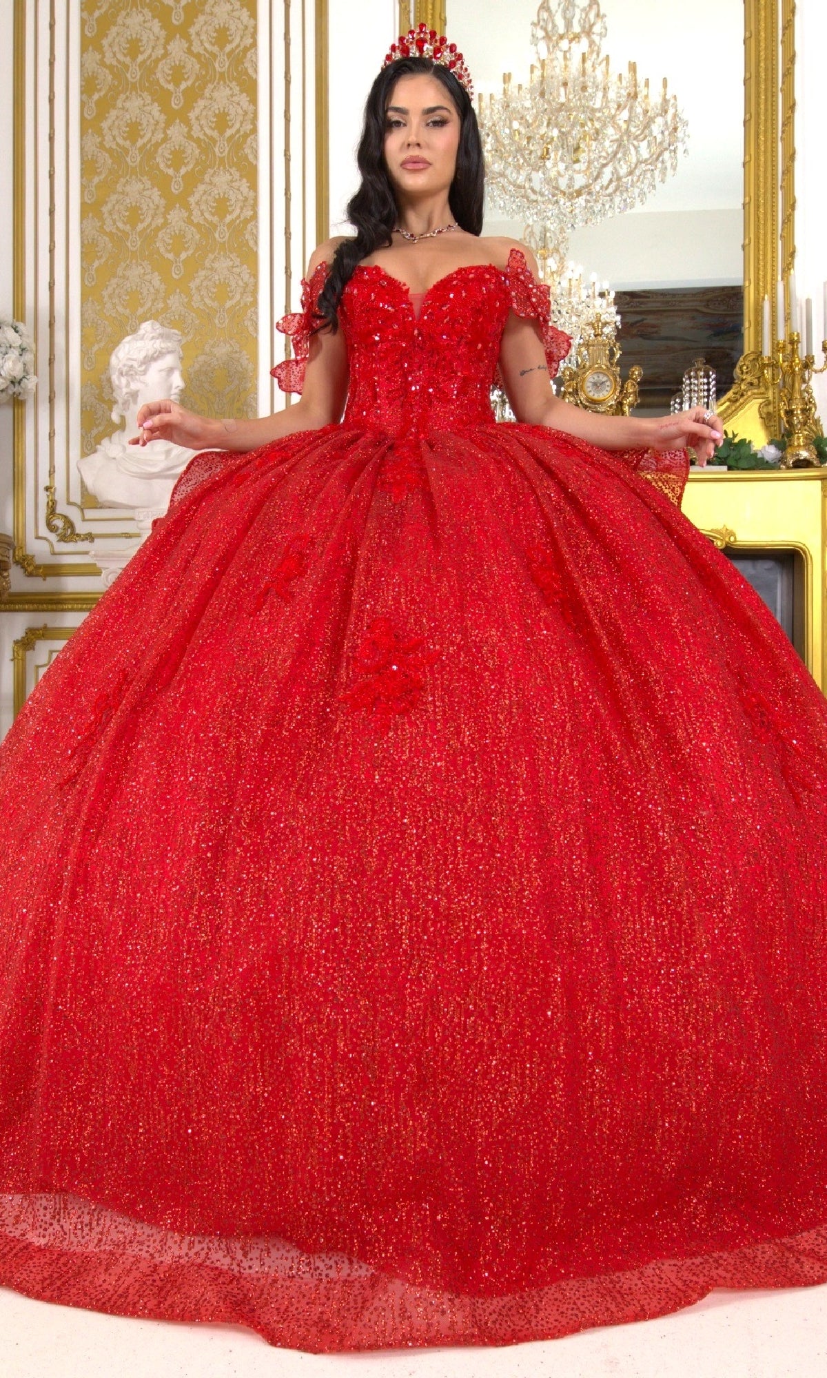 A woman stands in an ornate room wearing the Quinceanera Ball Gown 8120J, a sparkling red off-the-shoulder dress with lace sleeves, butterfly back detail, and a matching red crown.