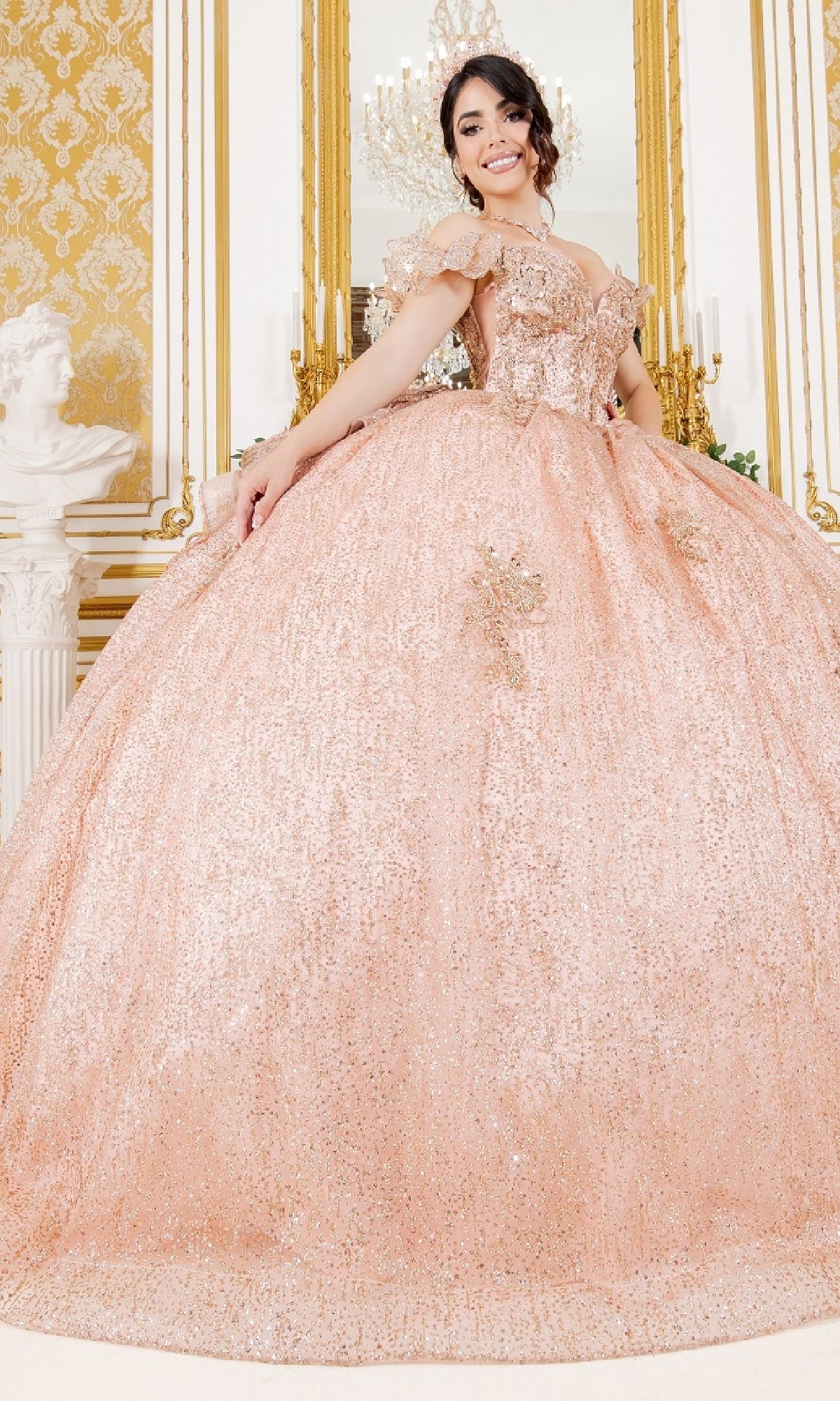 A young woman stands in an ornate room wearing the Quinceanera Ball Gown 8120J, a sparkling blush dress with floral accents, off-the-shoulder sleeves, and a delicate butterfly back detail.