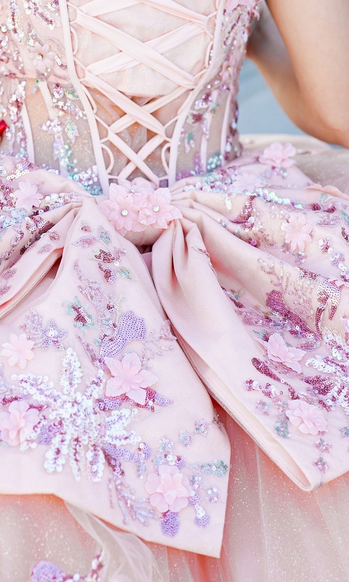 Close-up of Quinceanera Ball Gown 8125J, an off-the-shoulder light pink dress with a lace-up back, large bow, floral embroidery, sequins, and fabric flower accents.