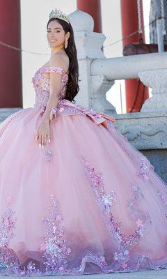 A young woman in the Quinceanera Ball Gown 8125J—an off-the-shoulder, pink floral dress—wears a tiara and poses outdoors before a stone railing and large red columns.
