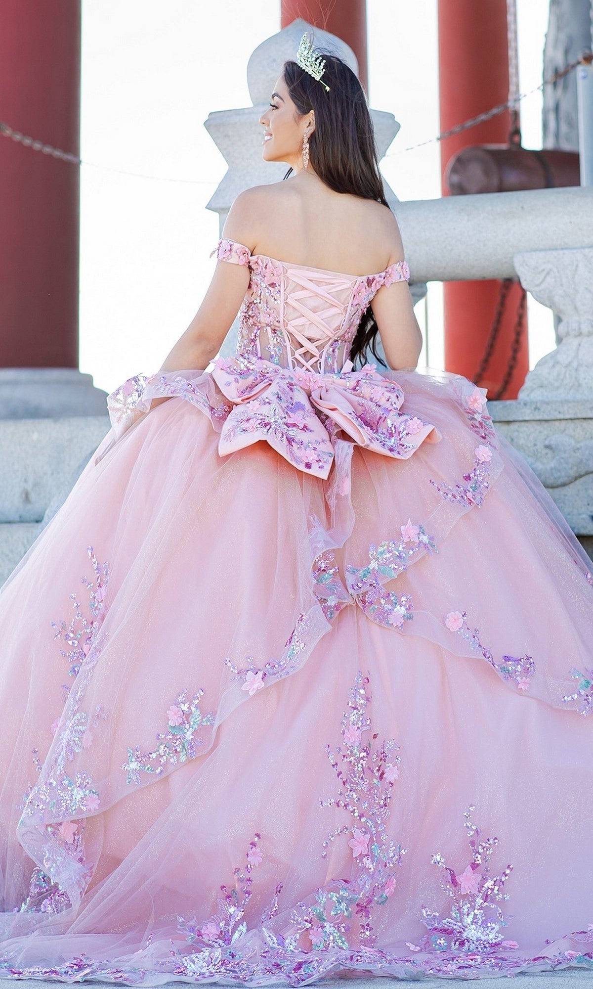 A young woman wearing the Quinceanera Ball Gown 8125J with a corset lace-up bodice and floral decorations stands on stone steps, tiara in place, facing away from the camera.