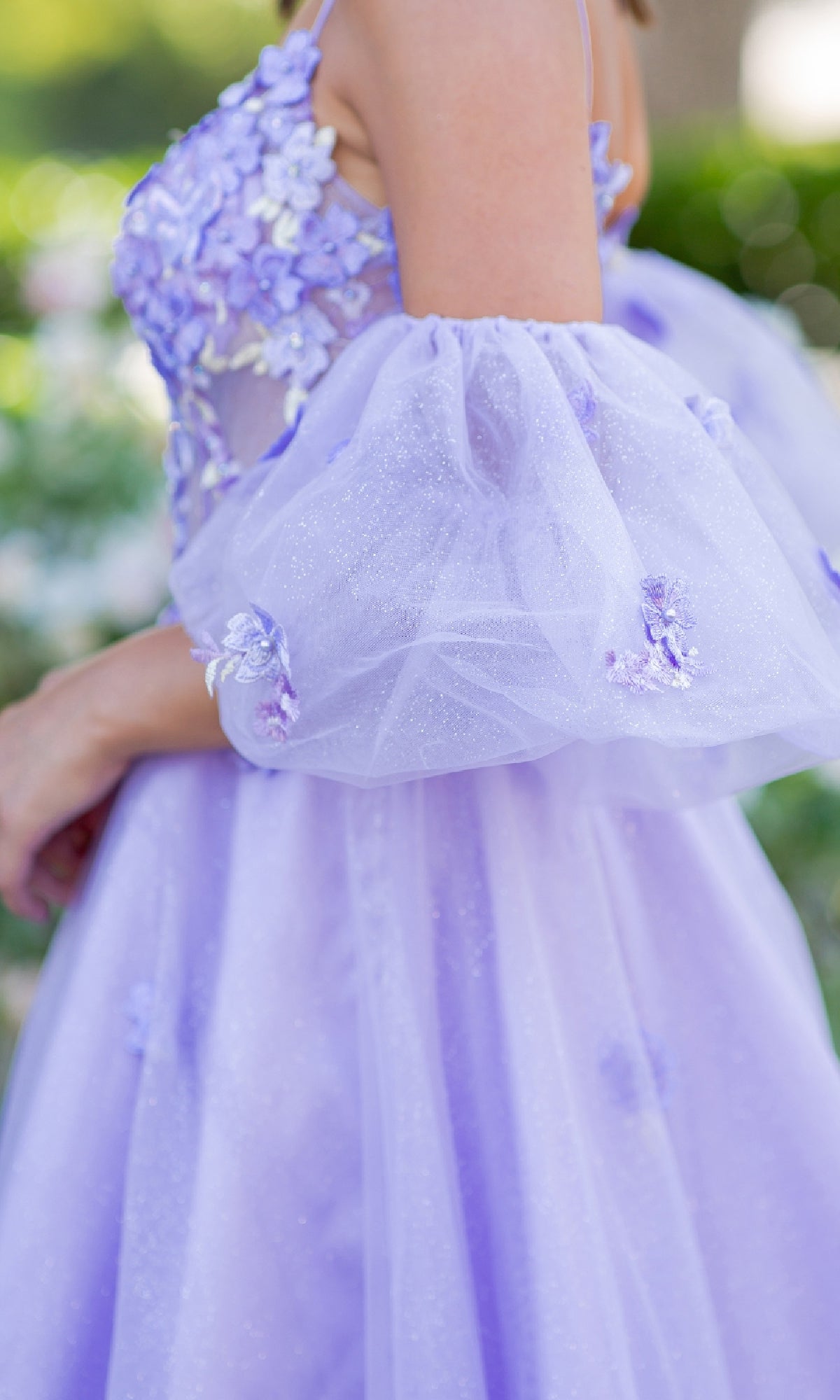 Someone stands outdoors wearing the Purple Prom Ball Gown: Cinderella Couture 8130J, featuring a lilac hue with floral embellishments and sheer, puffy sleeves.