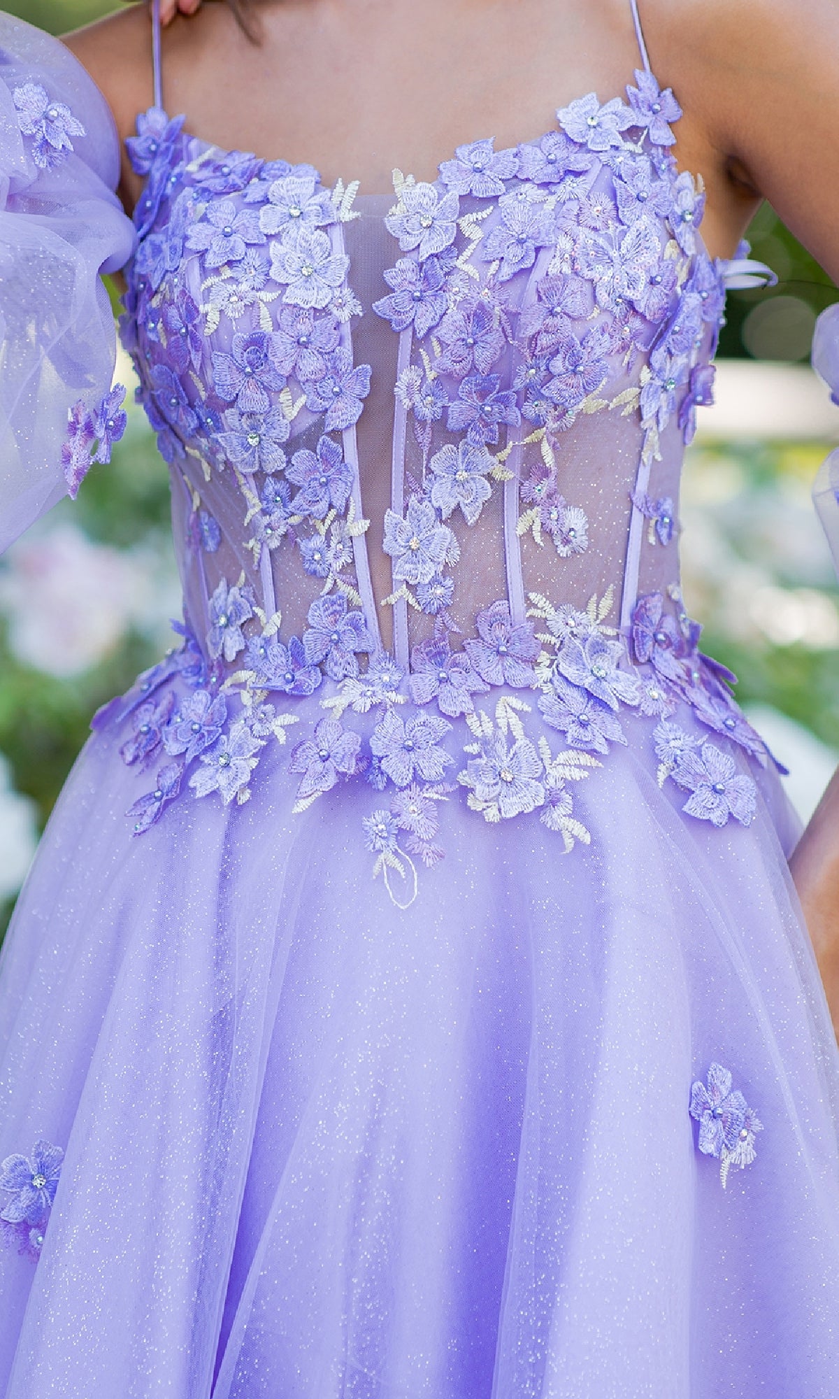 A close-up of the Cinderella Couture 8130J Purple Prom Ball Gown, featuring sheer panels, floral appliqué, glitter accents, and thin straps, worn by a person outdoors.