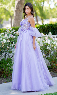 A woman stands outdoors in the Cinderella Couture 8130J Purple Prom Ball Gown, featuring floral appliqué and a sheer bodice, surrounded by greenery and white flowers.