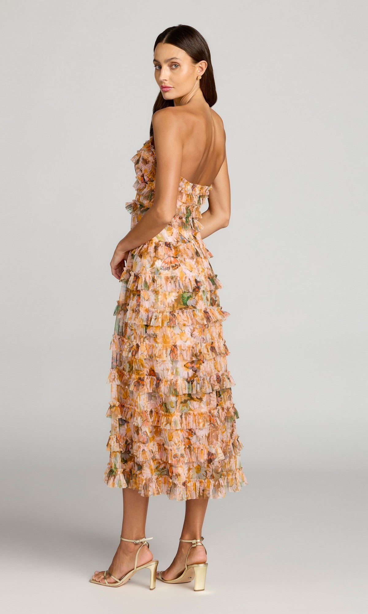 A woman stands in profile against a plain gray background wearing the Mac Duggal 8137 Strapless Long Print Formal Dress with layered floral ruffles and an orange pattern, paired with beige heeled sandals.